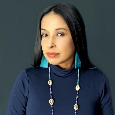 Headshot of the Latinx poet Peggy Robles-Alvarado wearing a sacred beaded necklace of cowrie shells with dark and light blue beads and large light blue rectangular earrings.